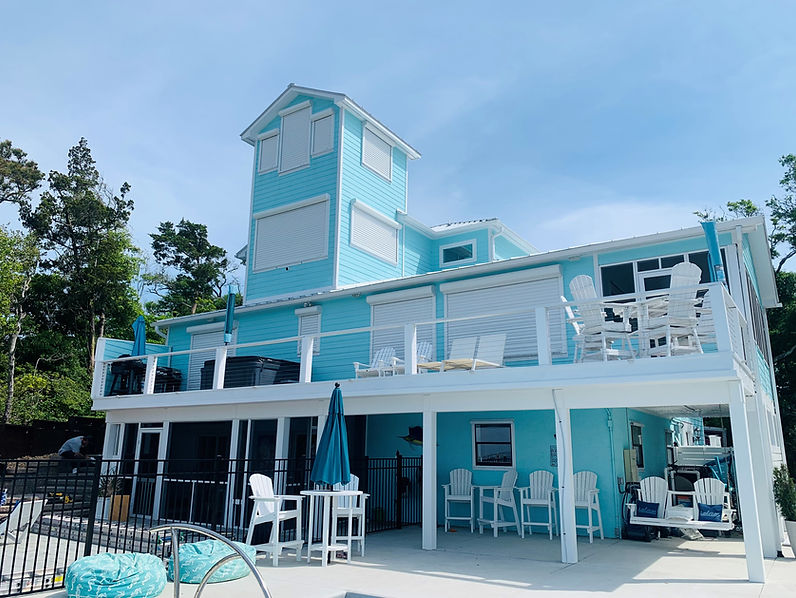 Atlantic Breeze Storm Shutters: A large turquoise beach house with multiple decks, white railings, and outdoor seating under a clear sky.