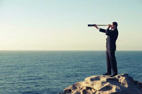 A man in a suit stands on rocks by the sea, looking through a telescope toward the horizon.
