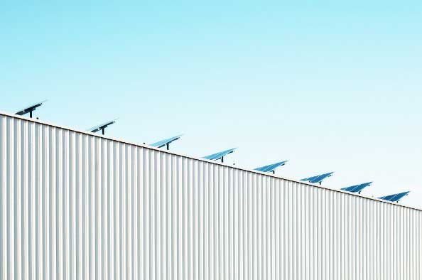 Solar panels are installed in a row on the roof of a large, white industrial building under a clear sky.