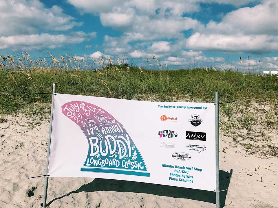 A banner on a sandy beach announces the 17th Annual Buddy Longboard Classic under a blue sky with clouds.