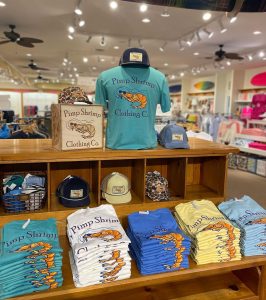 Display of colorful "Pimp Shrimp Clothing Co." shirts and hats neatly arranged in a bright retail store.