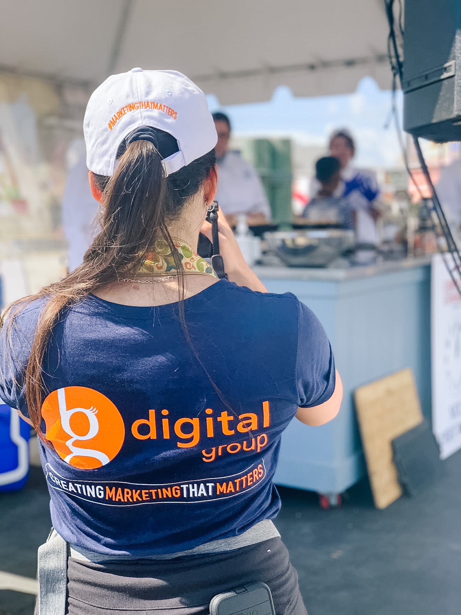 Woman wearing a "digital group" shirt takes a photo at an outdoor event under a white tent.