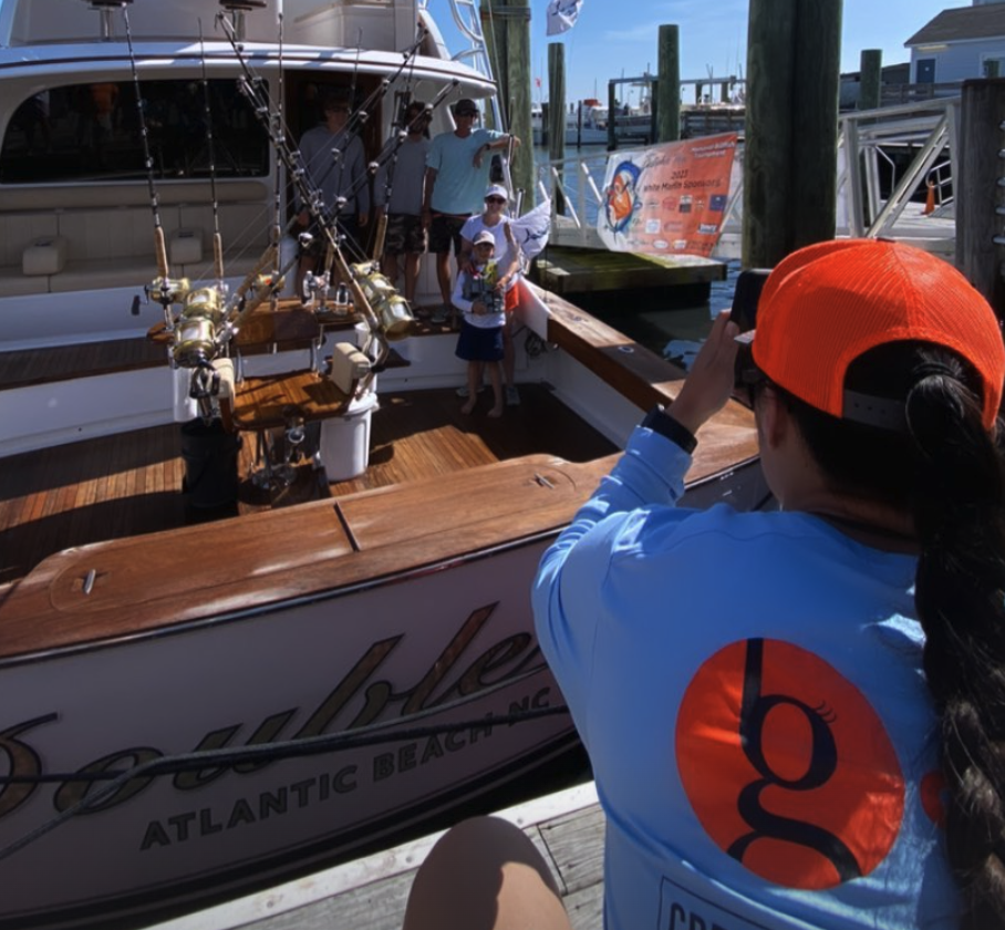 Person in blue shirt takes photo of two kids and two adults on a docked fishing boat in bright sunlight.