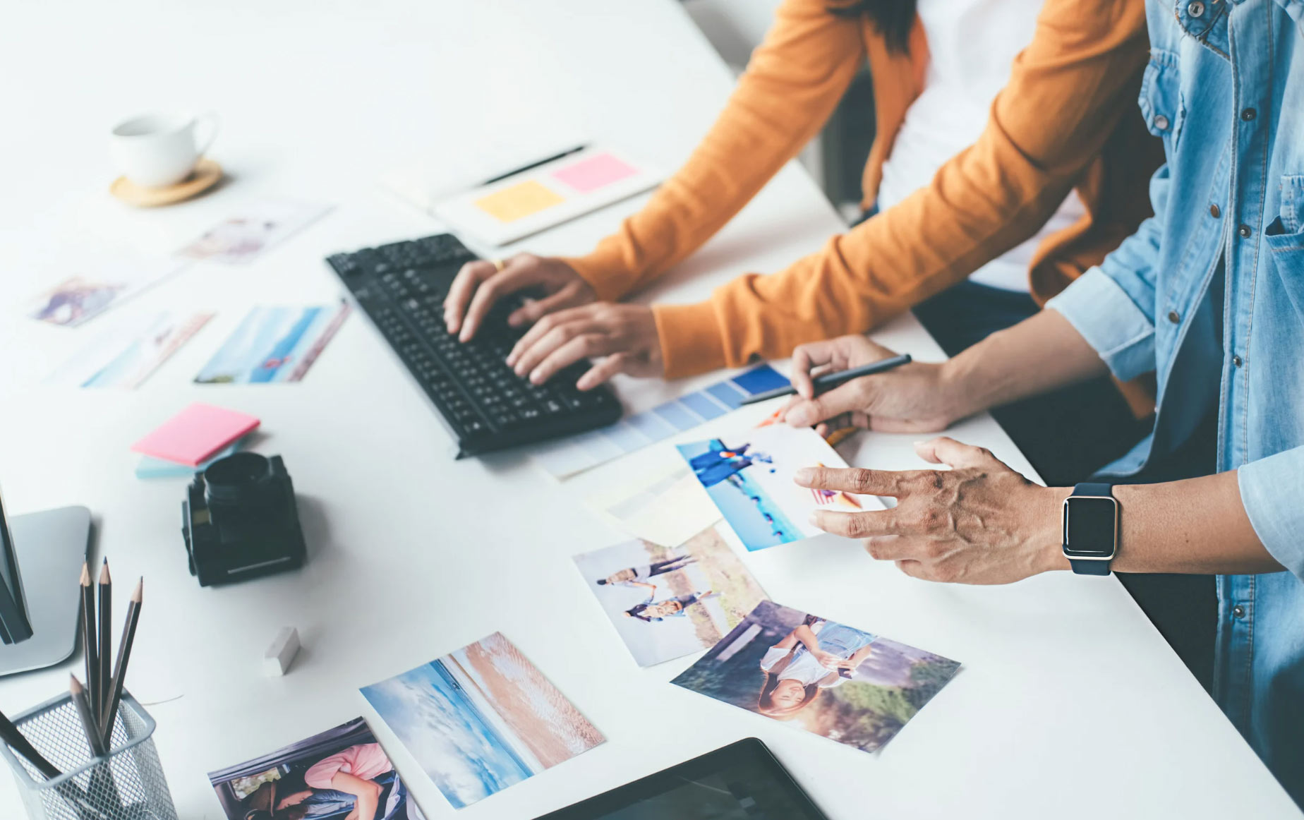 Two people editing and sorting photographs at a desk with a keyboard, camera, and photos spread out.