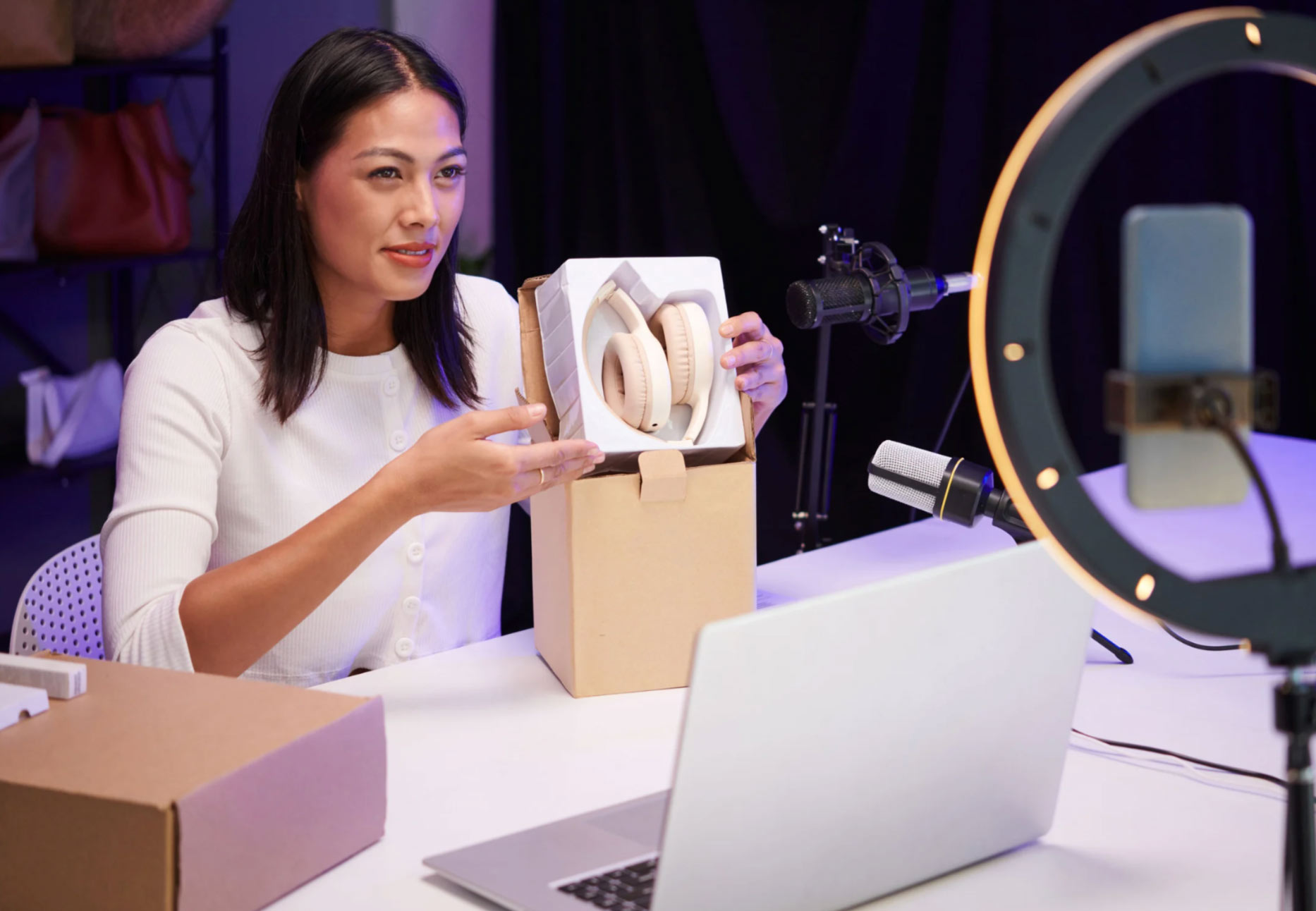 Woman unboxing headphones on camera at a desk with a laptop, microphone, and ring light.