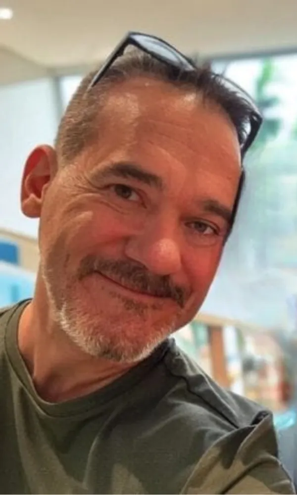 Smiling man with arms crossed, wearing a navy shirt, standing outdoors by the water.