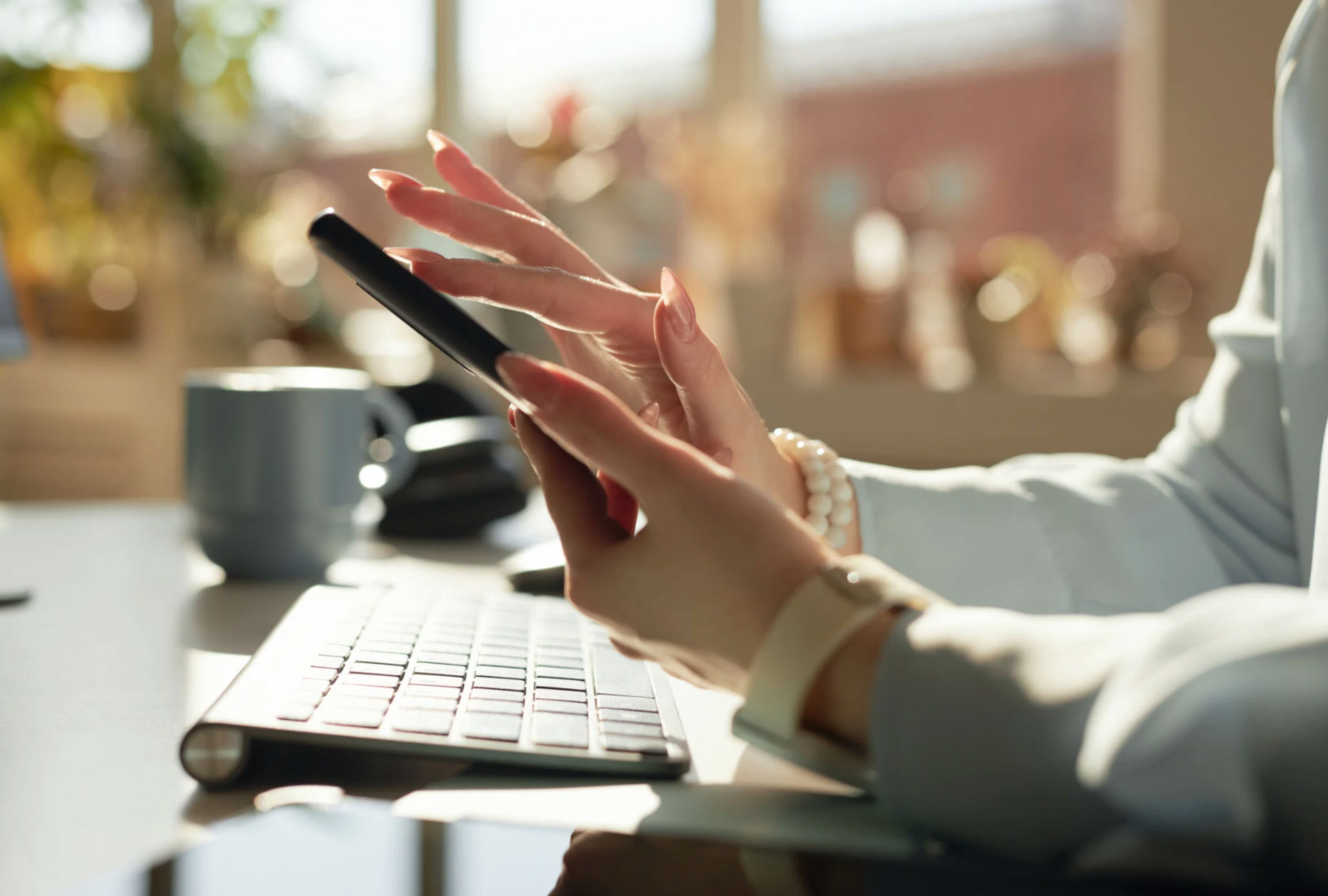 A person uses a smartphone at a desk with a keyboard, mug, and sunlight streaming through a window.