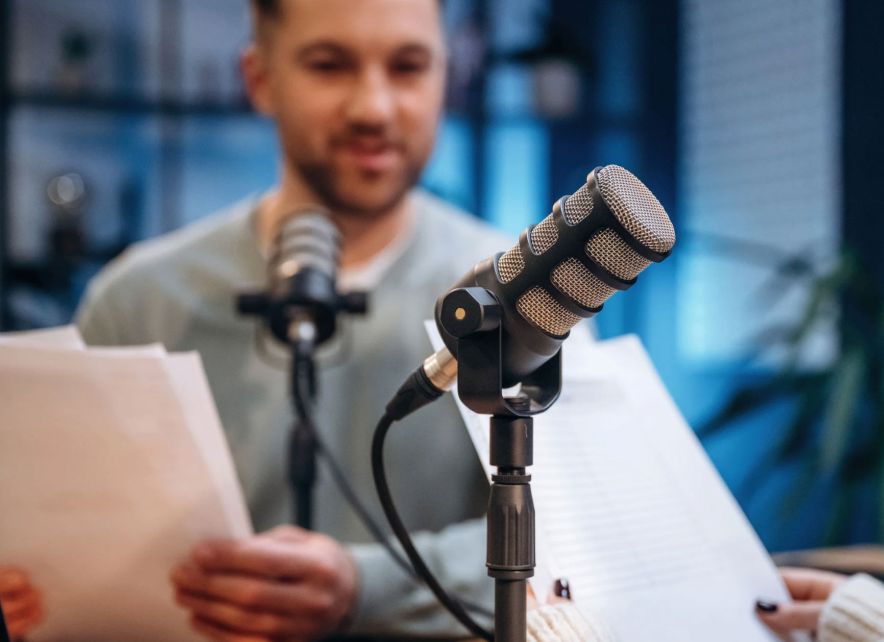Two people recording a podcast, holding papers, with microphones in focus in a studio setting.