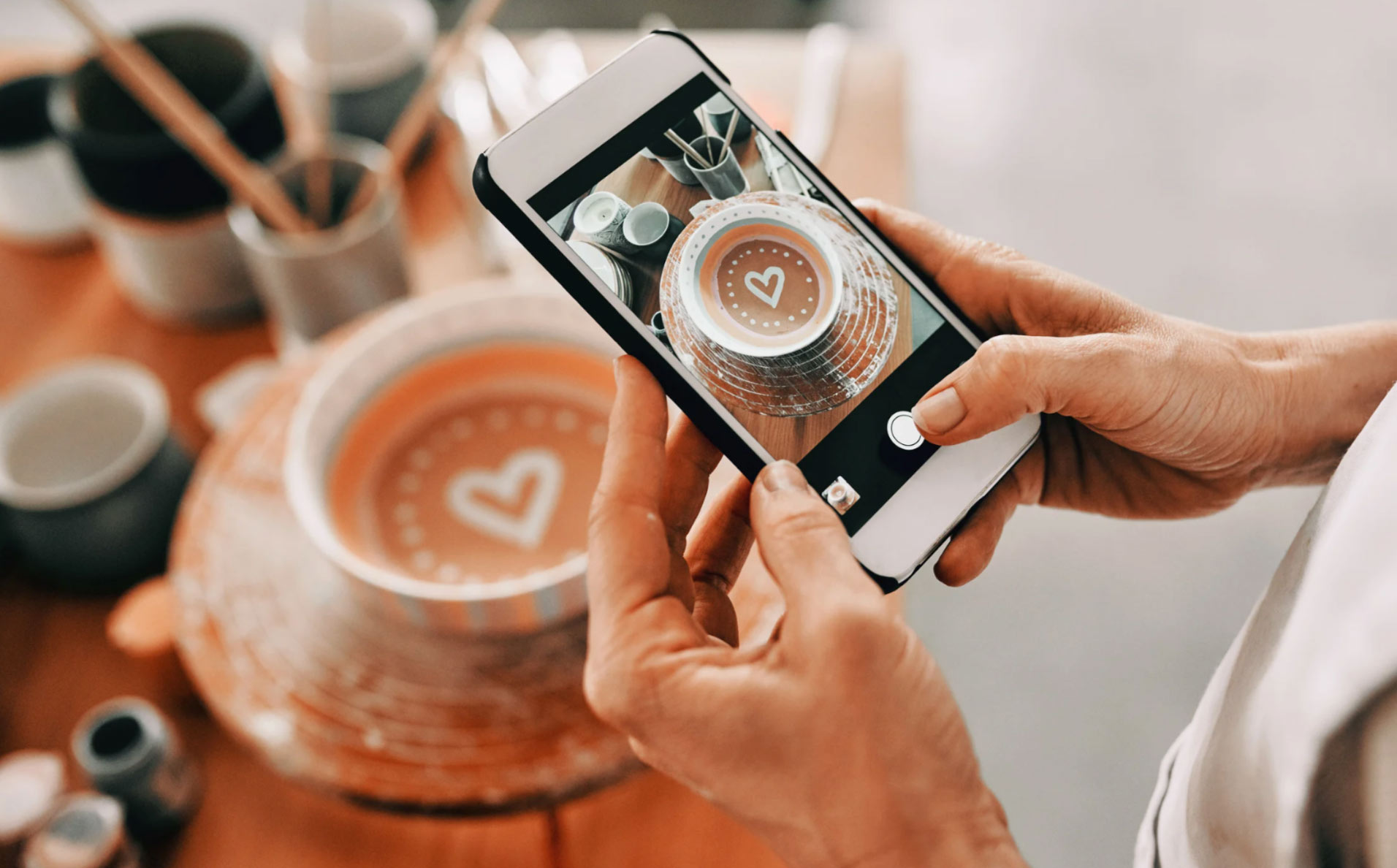 Person taking a photo of a heart-decorated ceramic bowl with a smartphone in a pottery studio.