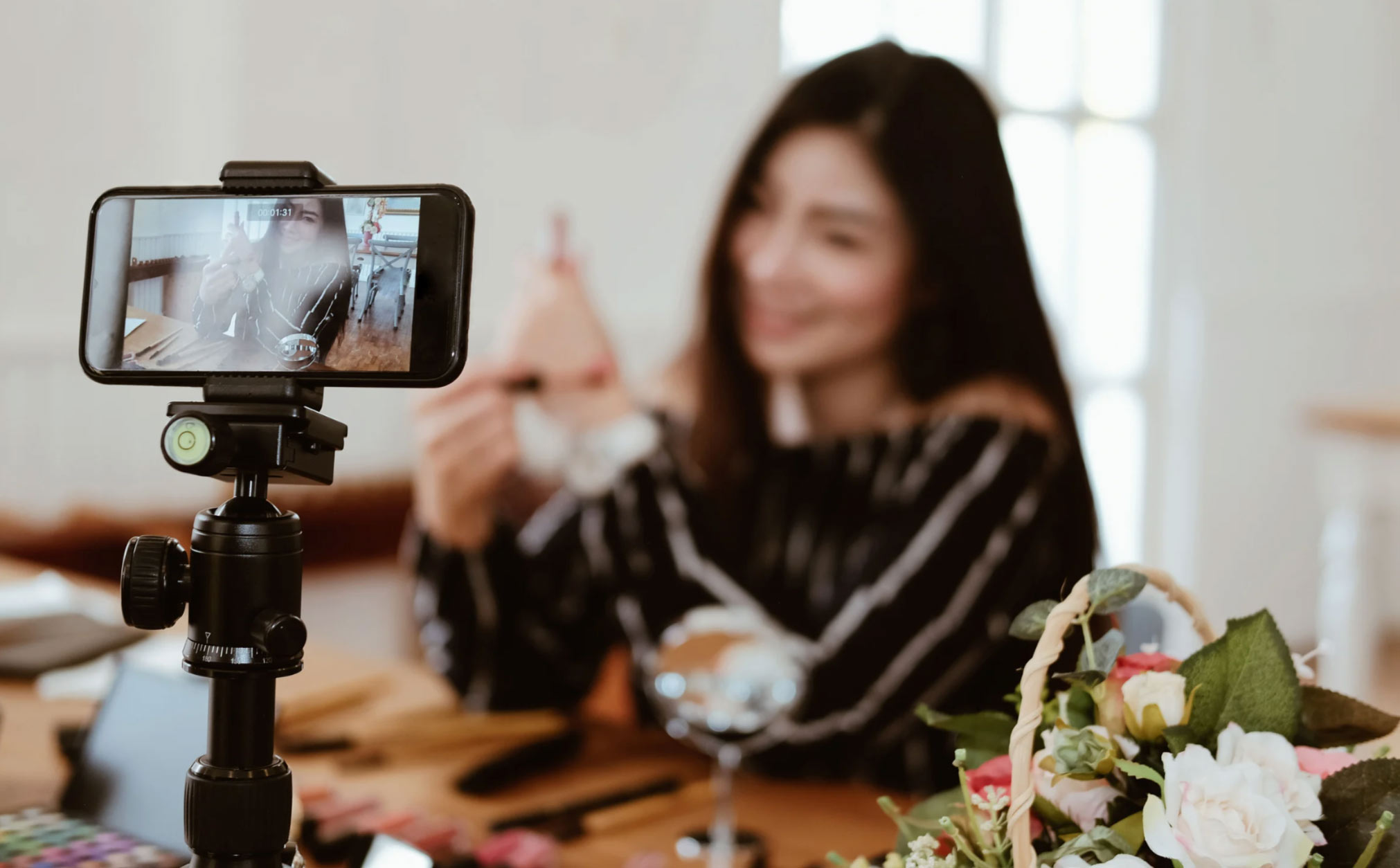 A woman livestreams from home, demonstrating an item with her phone mounted on a tripod.