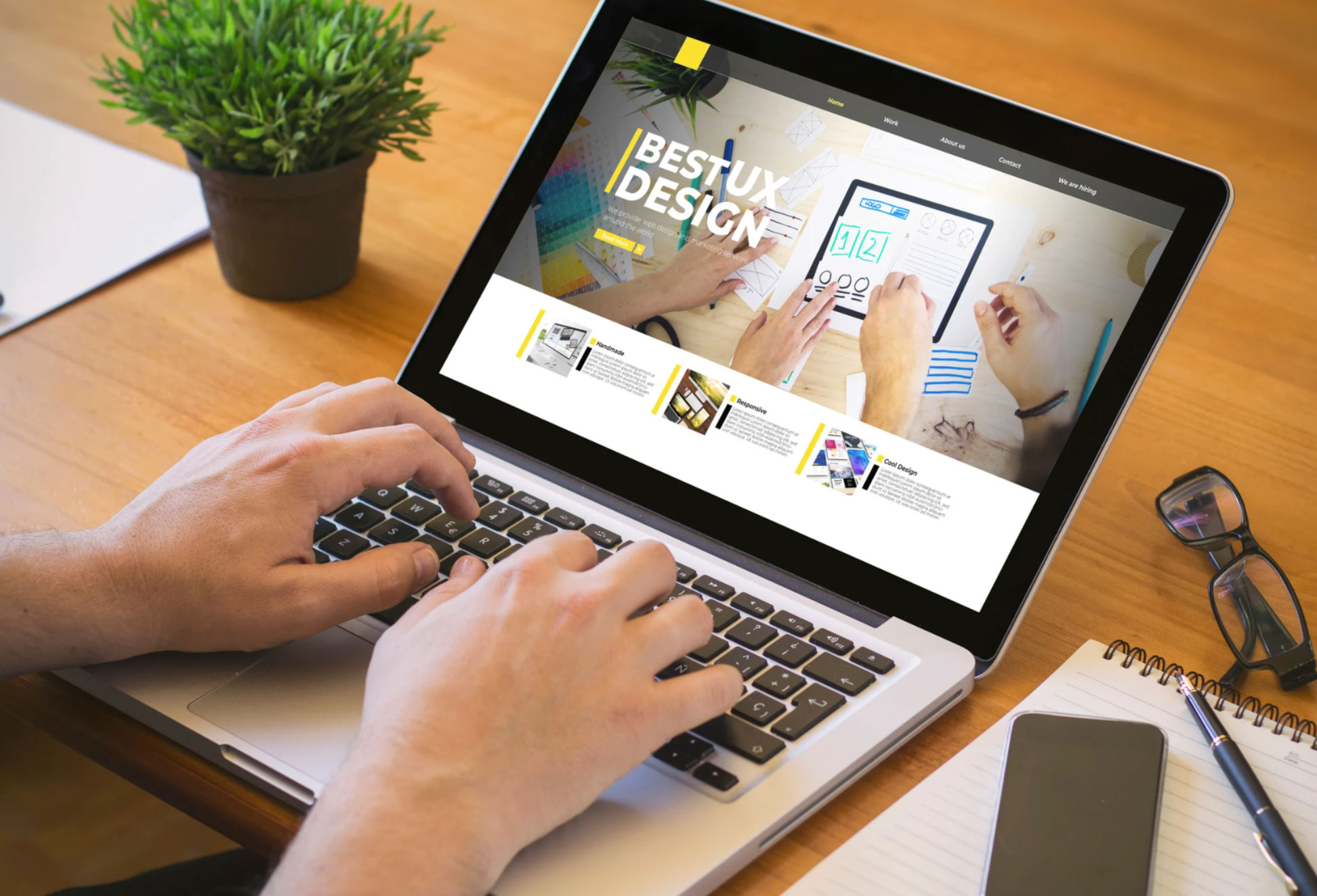 Person using a laptop displaying a "Best UX Design" webpage, with a notebook and plant on the desk.