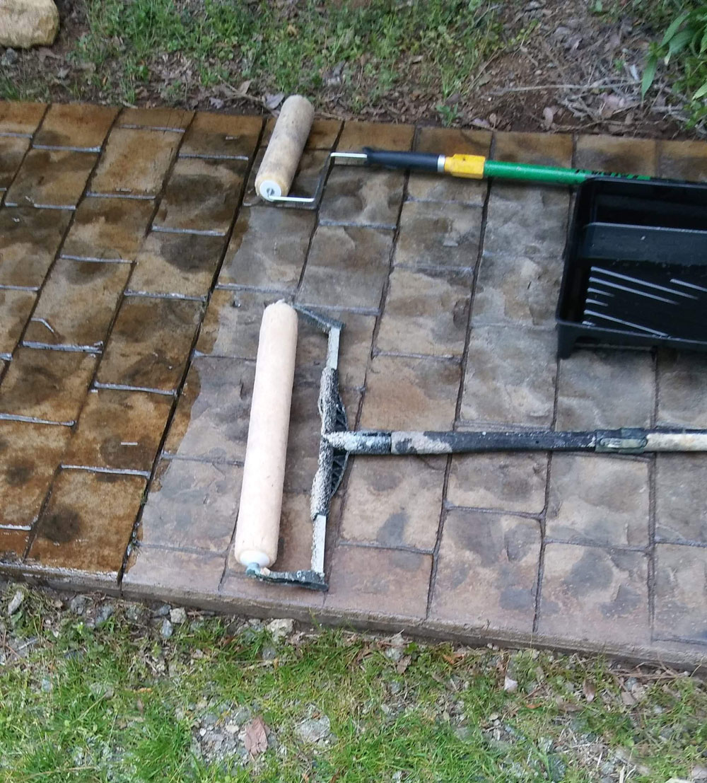 Paint rollers and a tray lie on a patterned stone path, part of which appears freshly coated or stained.