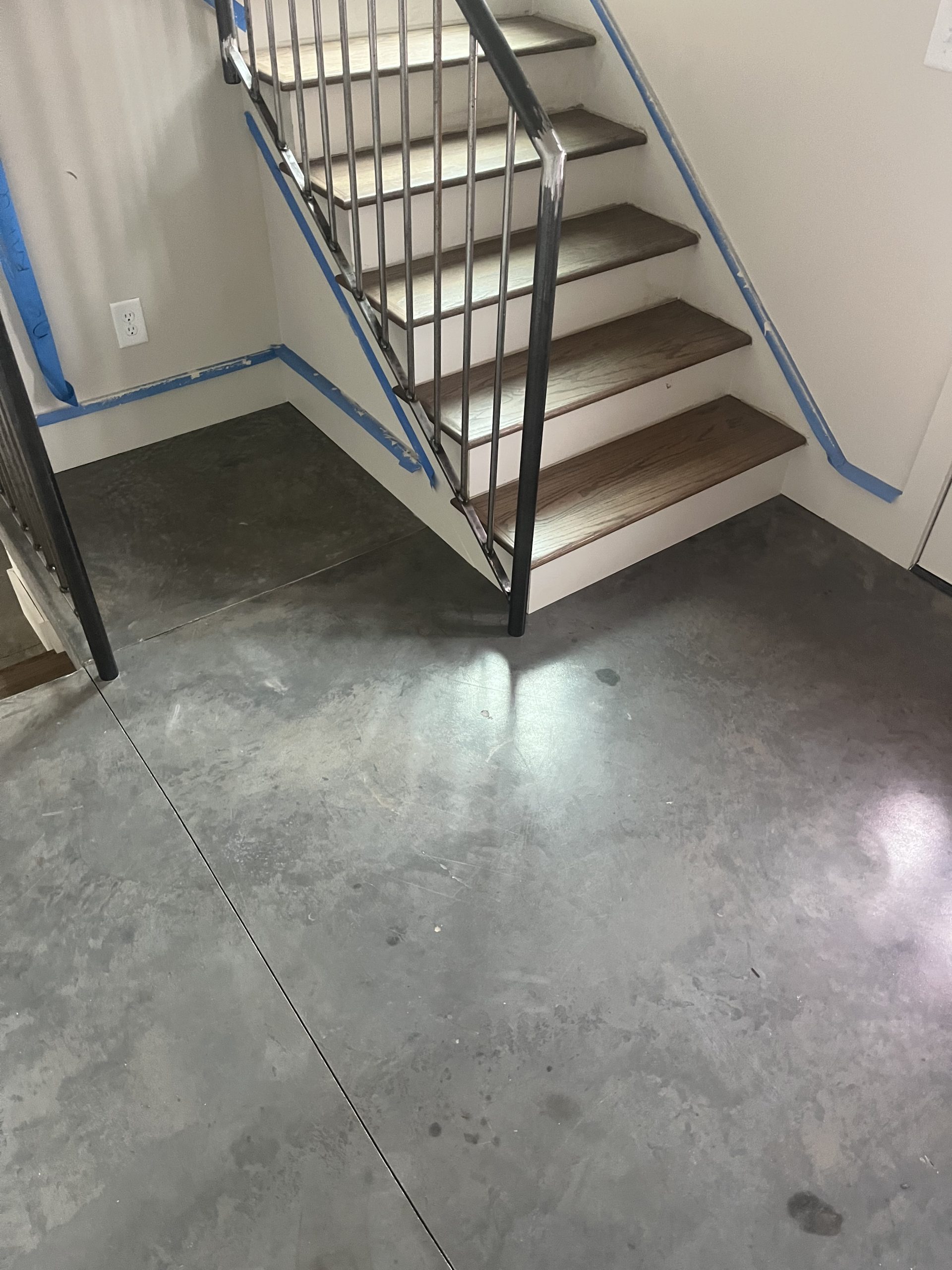 Stairs with wooden steps and metal railing, blue painter’s tape along walls and baseboards, concrete floor.