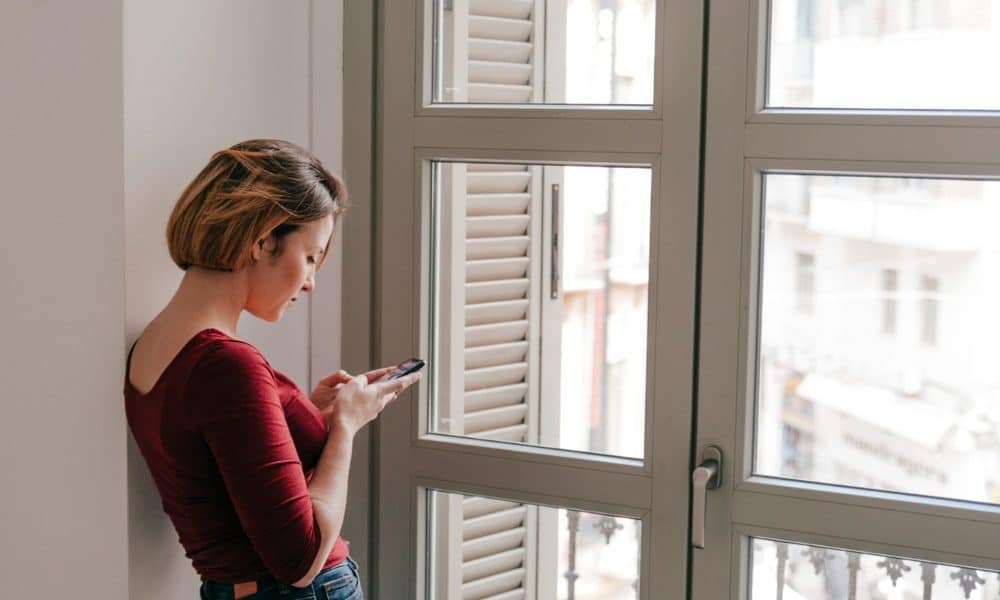 A woman stands by a large window with unique design elements, leaning against the wall as she texts on her phone.